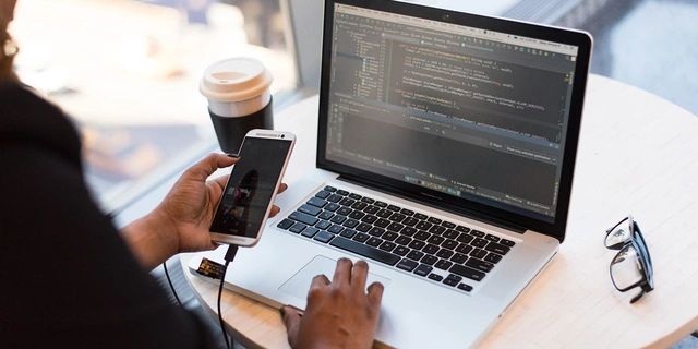 Person coding on a laptop while holding a smartphone.