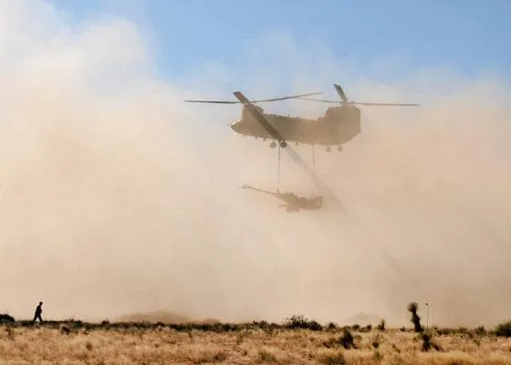 A military helicopter transporting a piece of equipment in the desert.