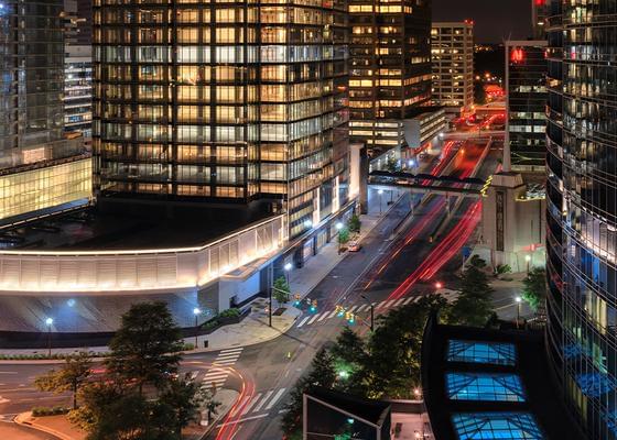 A photo of an urban setting at night with several tall modern building, and a multi lane intersection and road.